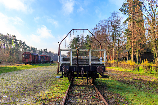 Old train station, flat car with coupling stops or bumpers on disused tracks, freight cars in background, bare trees against blue sky, sunny autumn day in As, Limburg, Belgium