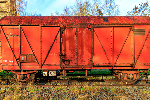 Rusty red metal freight car on disused train tracks at old station, bare trees against blue sky in background, sunny autumn day in As, Limburg Belgium
