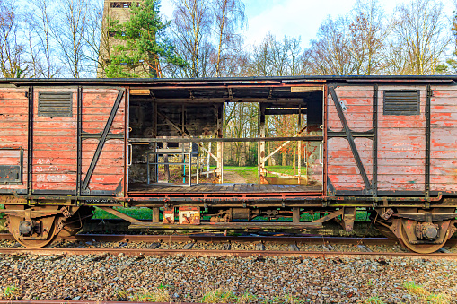 Damaged and broken wooden freight car on disused train tracks at old station, bare trees and part of observation tower in background, sunny autumn day in As, Limburg Belgium