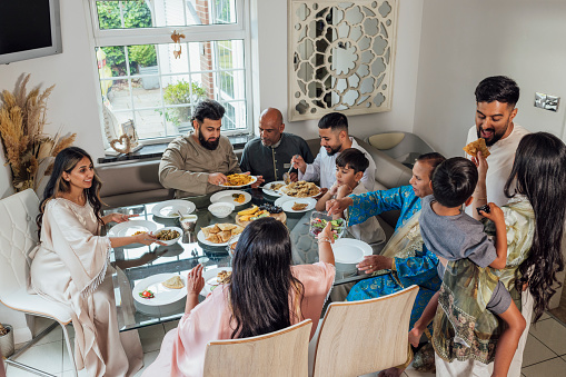 A multi-generational family gathered for Eid celebrations at home in Middlesbrough, North East England. They are all dressed in traditional outfits for the occasion. They are sitting around a dining table together where they share food and smiles.