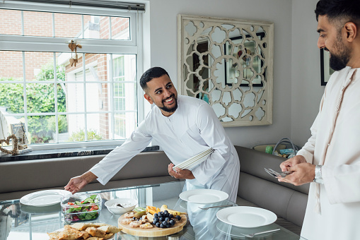 Two adult brothers gathered for Eid celebrations at home in Middlesbrough, North East England. They are all dressed in traditional outfits for the occasion. They are setting the dining table in the dining room.