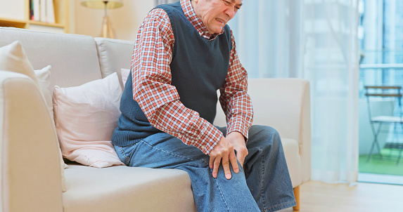 close up asian elderly man feel pain of knee sitting on sofa in living room at home - osteoporosis concept