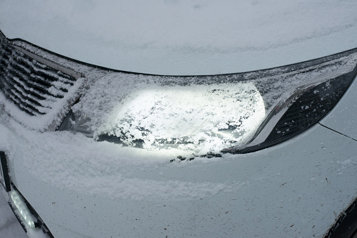 Close-up of the glowing headlights of a white modern car covered with snow in the evening in winter.