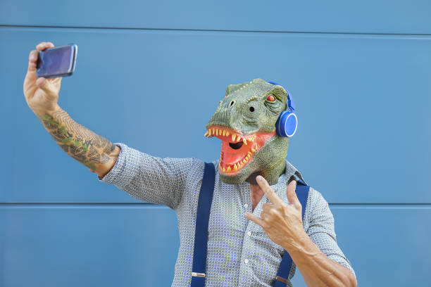 Crazy senior man wearing t-rex mask while taking selfie with mobile smartphone -  Hipster male having fun listening music and dancing outdoor -  Absurd surreal and technology concept