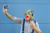 Crazy senior man wearing t-rex mask while taking selfie with mobile smartphone -  Hipster male having fun listening music and dancing outdoor -  Absurd surreal and technology concept
