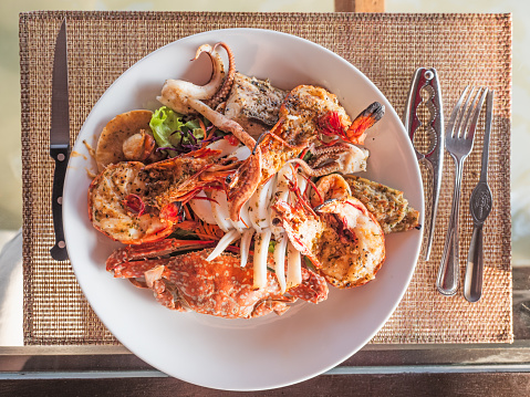 Seafood platter with grilled langoustine, crab, shrimps, squid and salad on a white plate with cutlery.