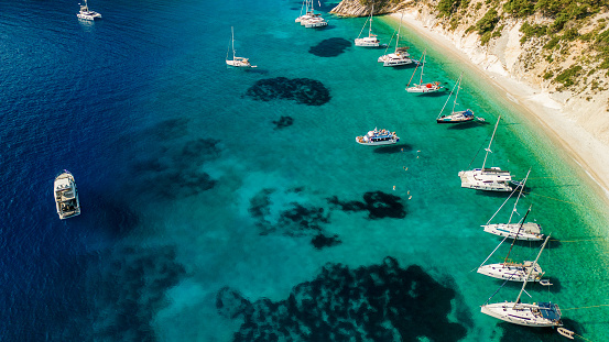 A beautiful bay with sailboats and catamaran on island of Ithaca, Greece. There are a dozen sailboats and two catamarans anchored in the bay. The sea is crystal clear, blue and turquoise.