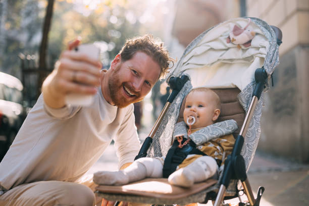 Embark on a journey through the chronicles of fatherhood as a ginger dad takes a selfie with his baby in the stroller, showcasing the love and happiness shared in this special moment captured on his mobile phone.