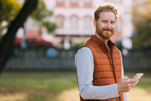 Escape into a world of technology amidst the beauty of nature as a ginger man, dressed in an orange vest, strolls through the park, seamlessly integrating his mobile phone into the outdoor experience.