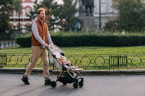 A ginger father, distinguished by his beard and orange vest, takes his baby for a delightful park promenade, pushing the stroller and radiating joy in this charming display of paternal pride.