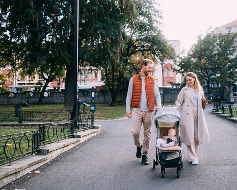 Embrace the peaceful ambiance of a family stroll in the park, where a mother and father leisurely walk, pushing their baby in a stroller, creating a harmonious and idyllic scene.