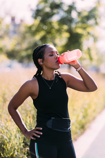 Elevate the running experience with water wellness as an African-American woman takes a moment to hydrate, enhancing her physical well-being amidst the park's beauty â a celebration of the symbiosis between runner and nature.