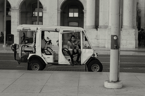 Lisbon, POrtugal - November 11, 2023: A tuk-tuk driver waits for clients while sittinhg in his tuk-tuk at the Praça do Comércio square in Lisbon downtown.