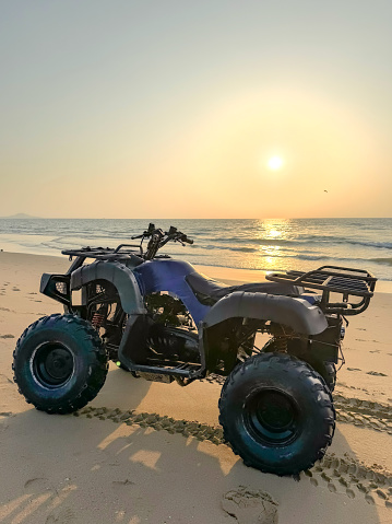 A serene moment captured at sunset, where a quad bike is parked on the sandy shores of a beach, with the golden sun setting over the calm waves in the background, painting the sky with warm hues.
