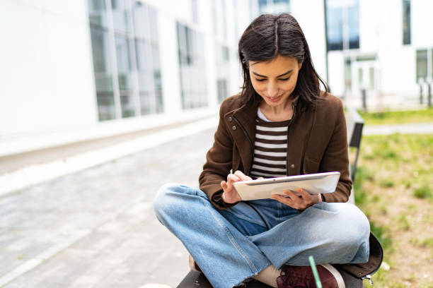 istock Female university student sitting on bench in campus using digital tablet 1919522457