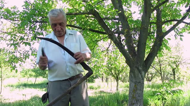 Senior Man Sharpening Scythe In The Garden