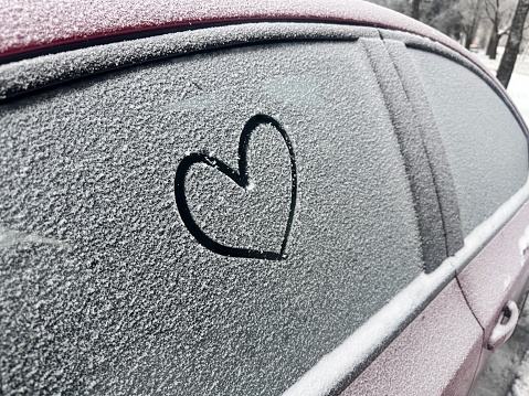Heart shaped doodle hand drawn on thin snow covered glass surface of the side window car in the winter season.