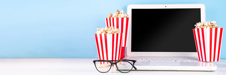 Cinema, film and movie night watching concept. Clapperboard, striped popcorn bucket boxes, with glasses and white laptop on light blue background, copy space