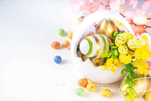 Cute Happy Easter spring holiday greeting card background. Colorful Easter eggs in basket with bunny rabbit decor, spring flowers bouquet on white wooden table background, copy space