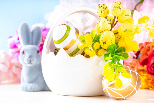 Cute Happy Easter spring holiday greeting card background. Colorful Easter eggs in basket with bunny rabbit decor, spring flowers bouquet on white wooden table background, copy space