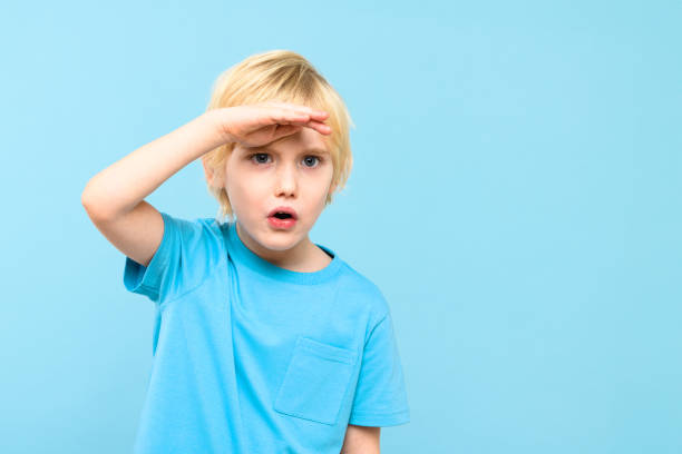 istock Cute preschooler looking curiously into distance with hand on the forehead. Kid looking into future, studio portrait on pastel blue background. 1919607537