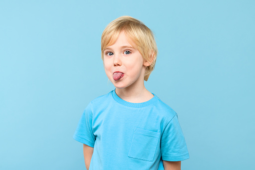 Funny young boy showing tongue and looking at the camera over pastel blue background. Cute preschooler grimacing at camera sticking out his tongue.