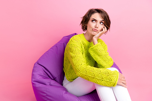 Photo of suspicious unsure girl dressed neon sweater sitting bean bag arm cheek looking empty space isolated pink color background.