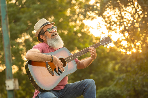 Ecstatic senior male playing guitar while spending leisure time in park during sunset