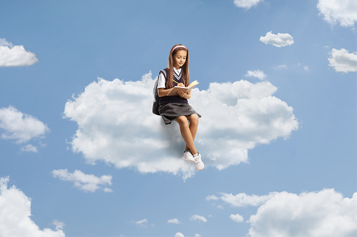 Girl in a school uniform sitting on a cloud and writing homework up in the sky