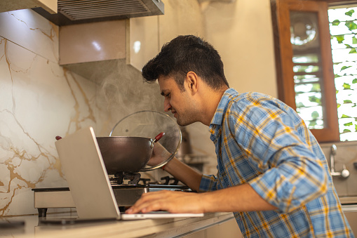 Man working over laptop on countertop and opening lid of utensil while cooking in kitchen at home