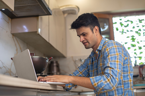 Man working over laptop on countertop while cooking in kitchen at home
