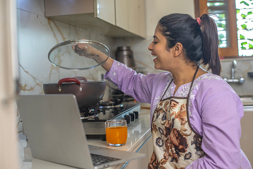 Businesswoman working over laptop on countertop and opening lid of utensil while cooking in kitchen at home