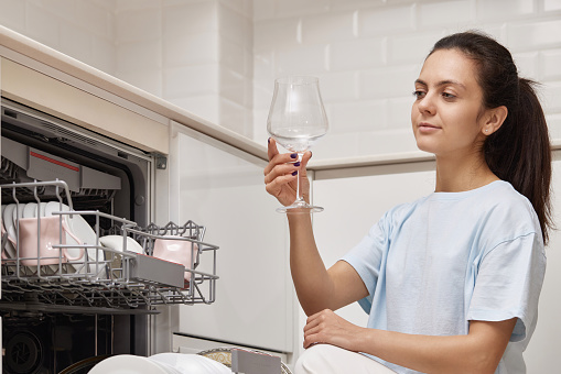 pretty woman takes wine glasses out of dishwasher and smiles in modern kitchen.