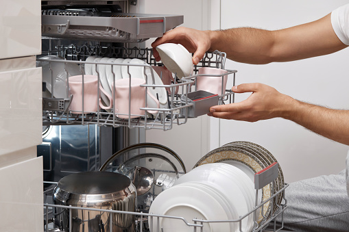male hand unloading plates from open automatic dishwasher machine with clean utensils inside in kitchen.