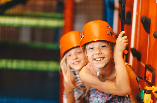 Two happy school girls in sport helmets at climbing class. Indoor bouldering class for school children. Kids enjoy at amusement park