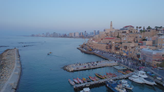 Jaffa port and the Old city at sunset with lots of families visiting restaurants, shops and bars in the port, Tel Aviv towers in the background - push in drone shot