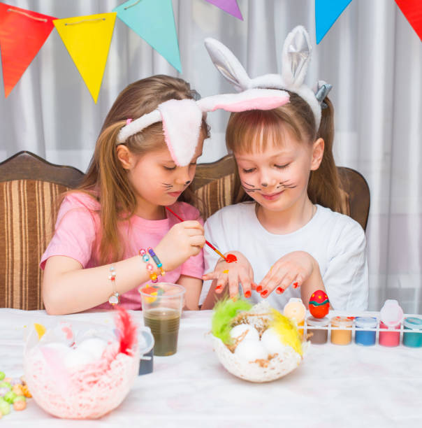 istock Girls in bunny ears having fun painting eggs and each other preparing for Easter. 1919962094