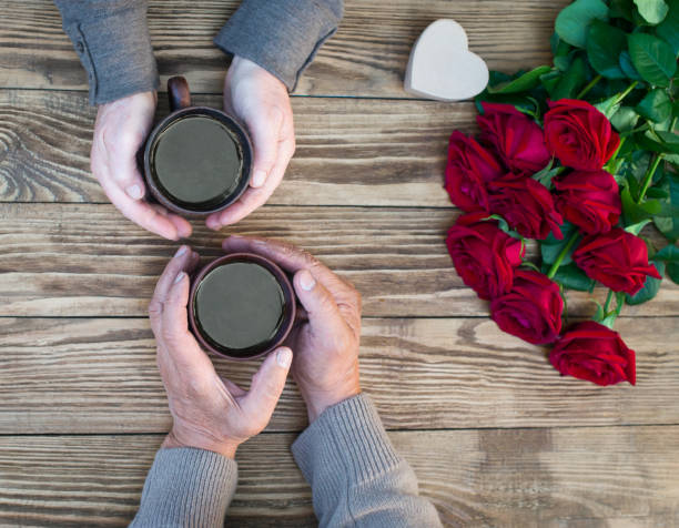 Senior couple drinking coffee and talking. Valentines background. Old people hands, coffee cups, roses, gift boxon wooden background.
