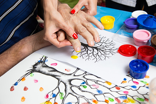Art therapy to restore finger motility in the hospital. The medical worker helps the grandfather to draw with prints