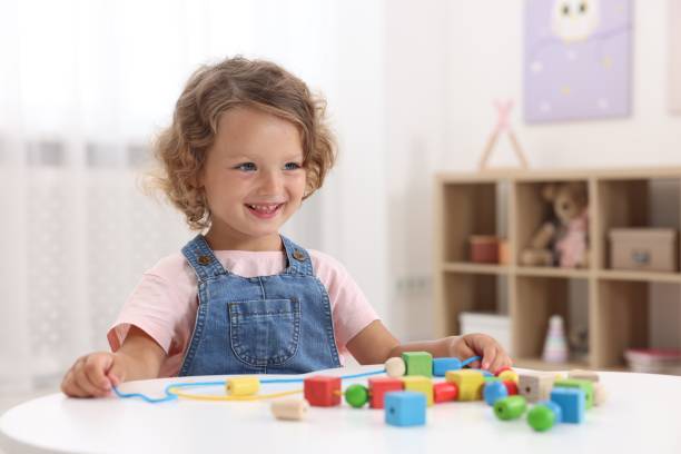istock Motor skills development. Little girl playing with wooden pieces and string for threading activity at table indoors 1920095344