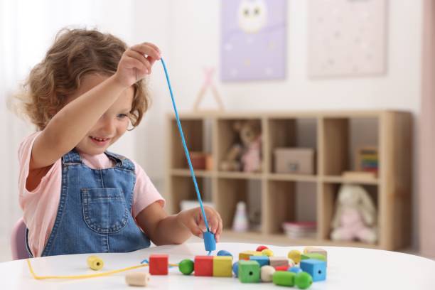 istock Motor skills development. Little girl playing with wooden pieces and string for threading activity at table indoors 1920095347