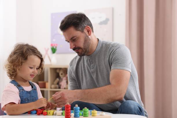istock Motor skills development. Father and daughter playing with wooden pieces and string for threading activity at table indoors 1920095914