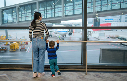 Waiting to board their flight, a mother and toddler standing by the window of an airport terminal and watch the plane. Active family lifestyle, travel with little child concept