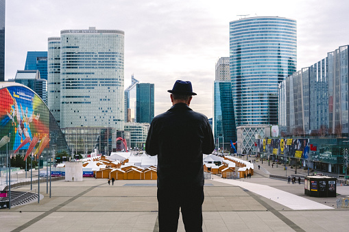 Paris, France - December 31, 2023: A man, with his back turned and wearing a hat, observes the business buildings of La Defense, in Paris.