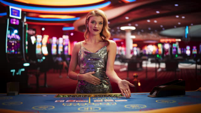 Beautiful Caucasian Woman Working As A Dealer At Classy Casino. Professional Female Baccarat Croupier In a Fancy Silver Dress, Presenting Playing Cards, Looking At Camera And Smiling In Slow Motion.