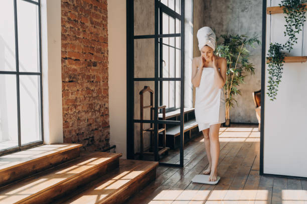 Woman in a spa towel stepping on a scale, looking down with a hopeful expression in a sunlit room with urban decor.