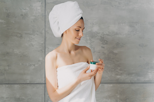 Serene woman wrapped in towels examining skincare product with a gentle smile in a minimalist spa setting