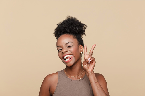 Headshot of excited black young woman wearing brown top standing against beige background, winking, sticking out tongue and showing victory sign.