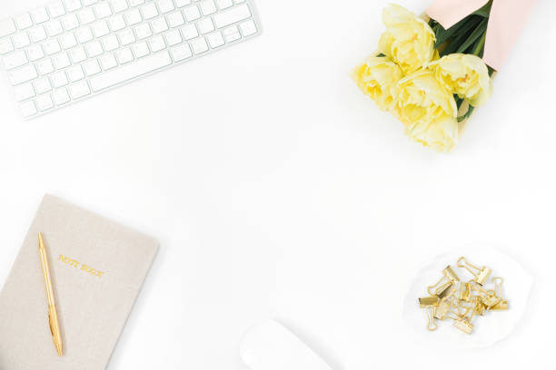 istock Stylized women's Desk, office Desk. A workspace with a computer, a bouquet of yellow tulips, a clipboard, a Notepad for notes and a ballpoint pen. Women's fashion accessories on a light white background. Flat top view 1920276652