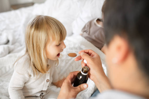 Father giving syrup to his sick little girl with flu, cold, coronavirus, bronchitis or pneumonia, virus or infection stock photo